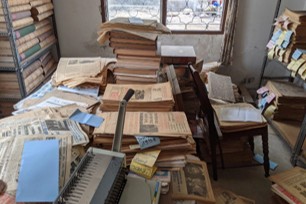 An unsorted backroom: stacks of old newspapers and bound volumes piled on tables, chairs and a sofa, with a scanner in the foreground — awaiting digitisation